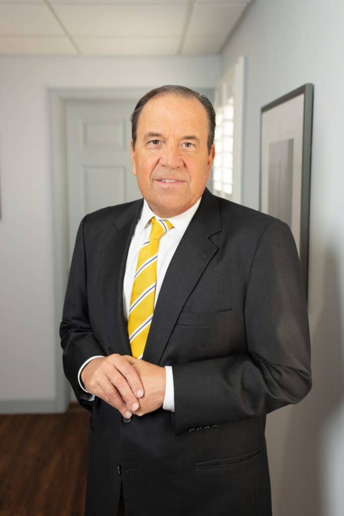 Professional portrait of attorney Lanny Darr in a law office setting. He is wearing a dark suit with a yellow striped tie, standing confidently with his hands clasped. The background shows a hallway and framed artwork, conveying a professional legal environment.