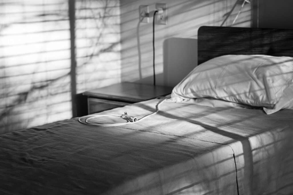 Empty hospital bed with a stethoscope lying on the sheets, in a dimly lit room with window blinds casting shadows. The black and white image evokes a sense of loneliness and potential neglect in a healthcare setting.
