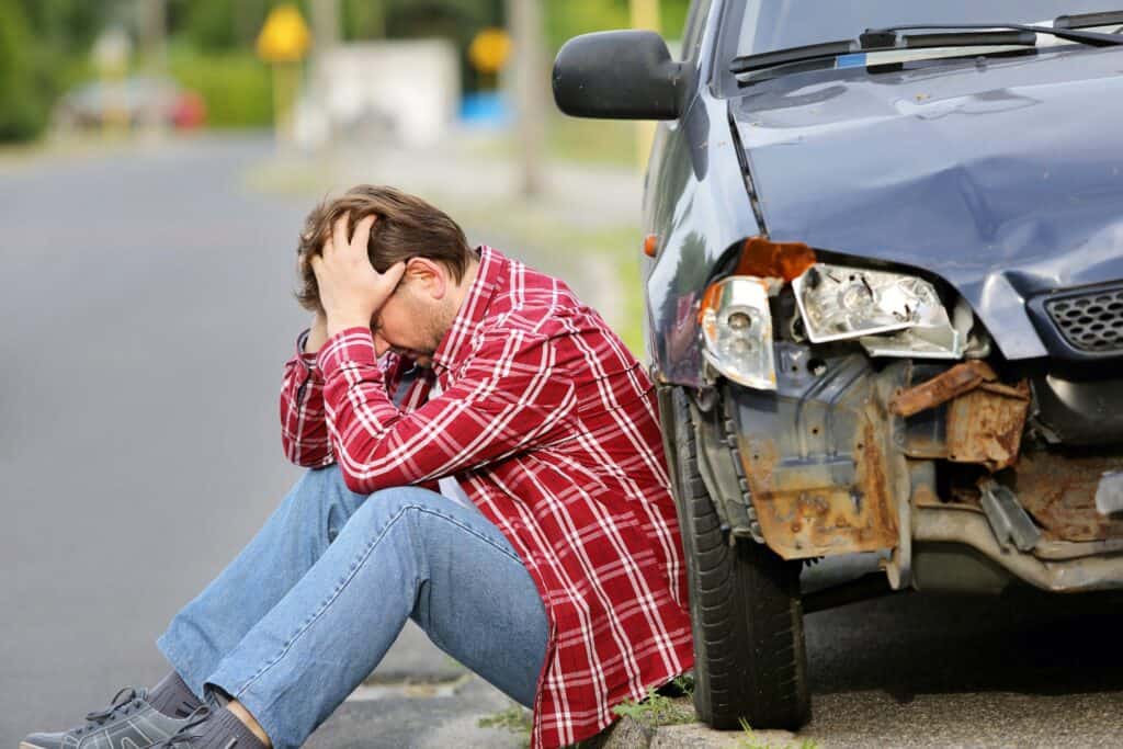 Distressed man in a red plaid shirt sitting on the ground next to his damaged car after an accident. The car's front end is visibly crushed, highlighting the impact of motor vehicle collisions.