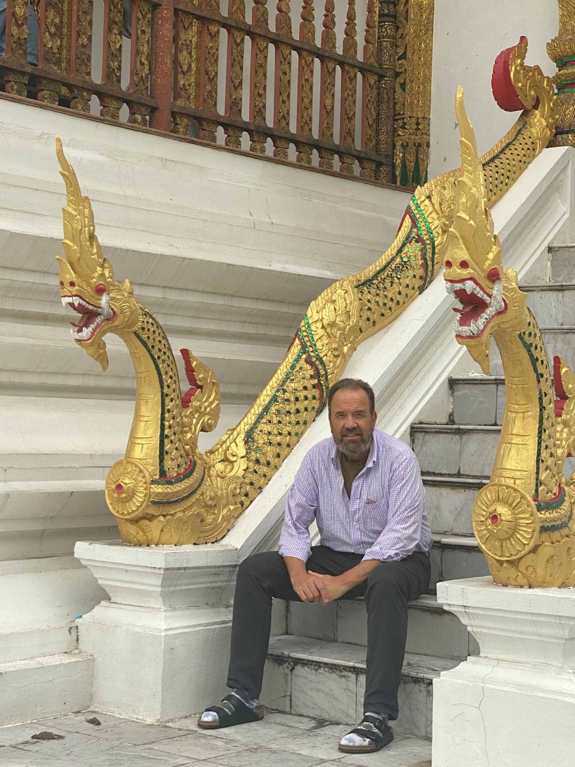 Lanny Darr sitting on the steps of an ornate structure with golden dragon sculptures, showcasing intricate design and cultural artistry.